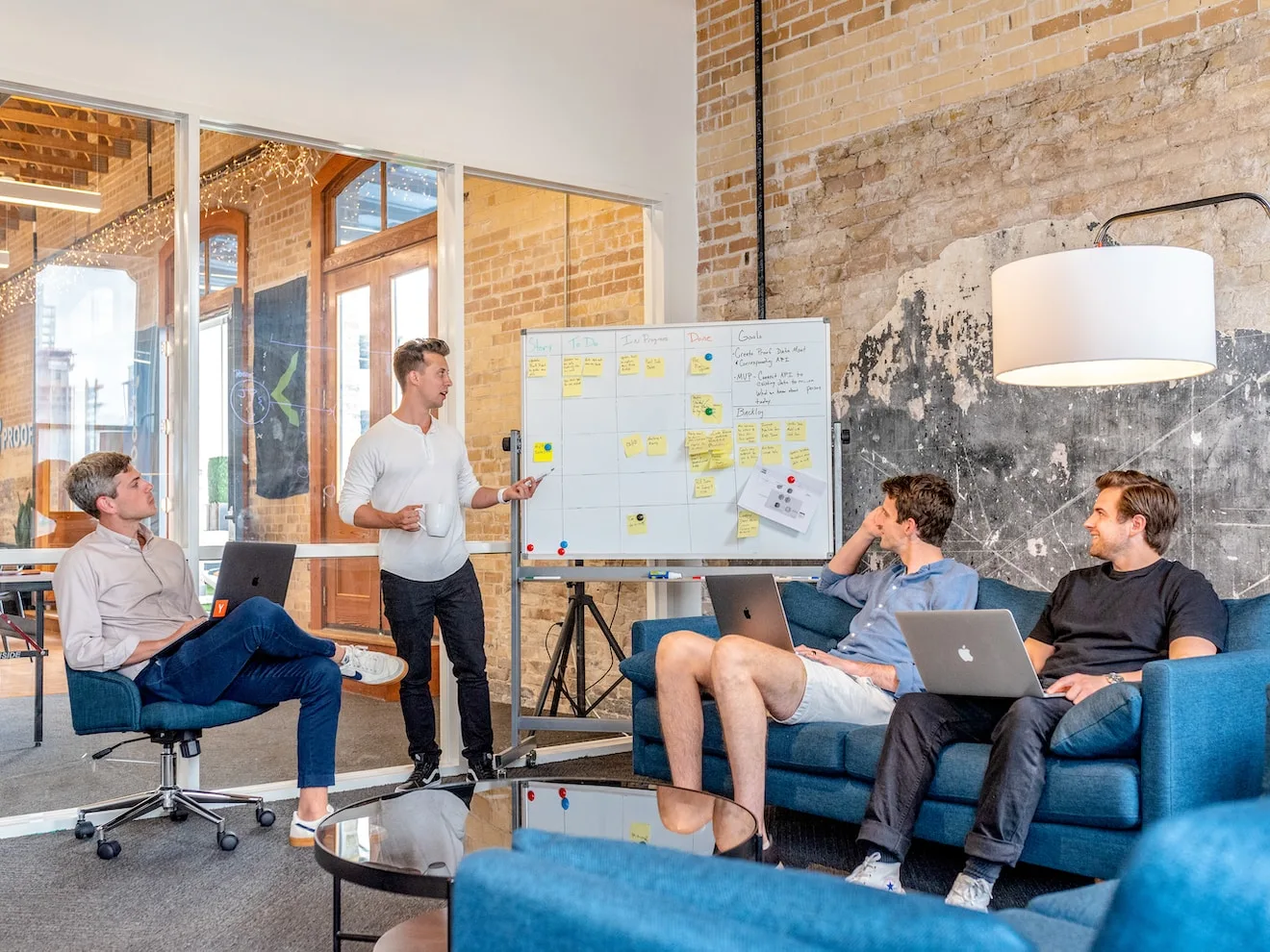 Team meeting in a brick-walled office, one person presenting at a whiteboard covered in sticky notes while three colleagues follow on laptops